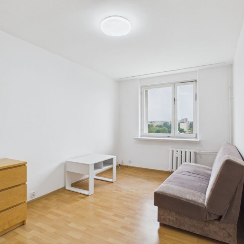 Sparse white living room with a brown sofa on the right, a small white coffee table, a wooden dresser on the left, and a large window with a radiator beneath; light wood floor throughout.