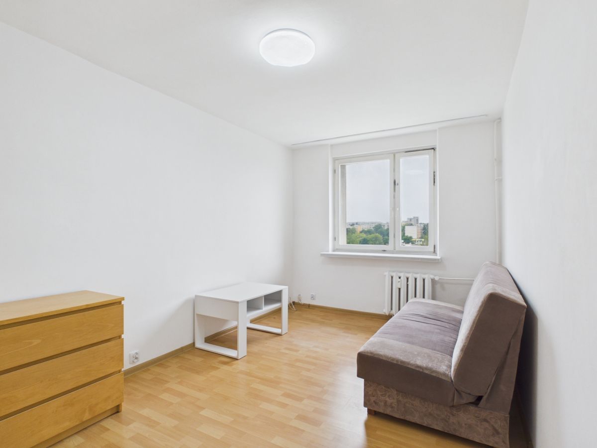Sparse white living room with a brown sofa on the right, a small white coffee table, a wooden dresser on the left, and a large window with a radiator beneath; light wood floor throughout.