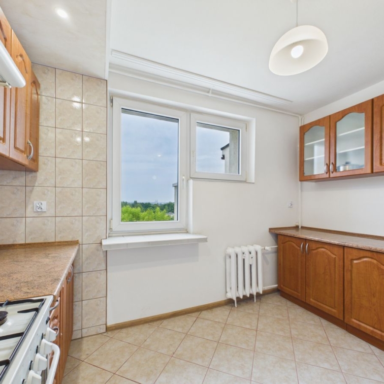 Bright kitchen with wooden cabinets, beige countertops, and tiled walls; gas stove visible on the left and a large window providing view of greenery outside.