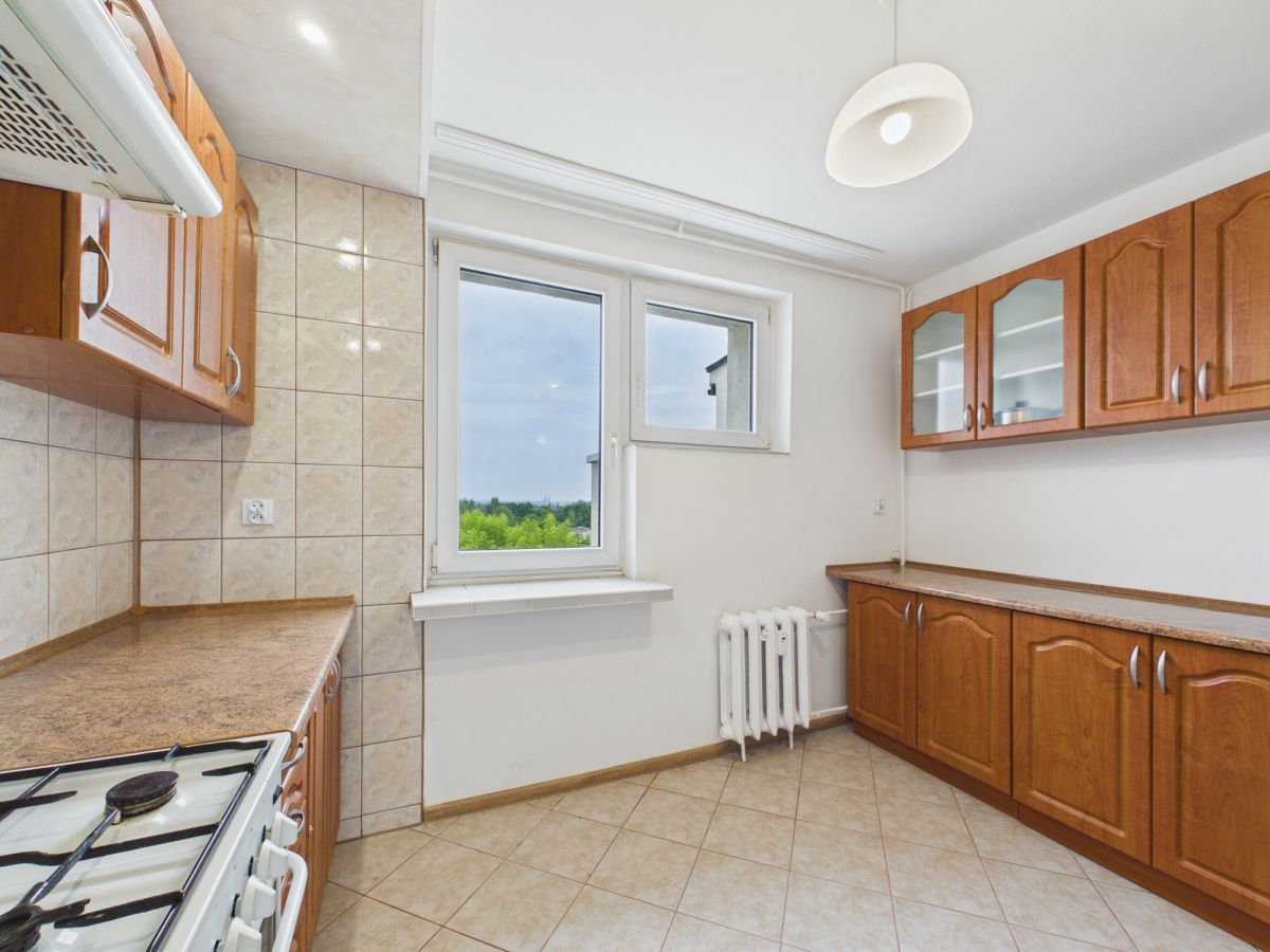 Bright kitchen with wooden cabinets, beige countertops, and tiled walls; gas stove visible on the left and a large window providing view of greenery outside.