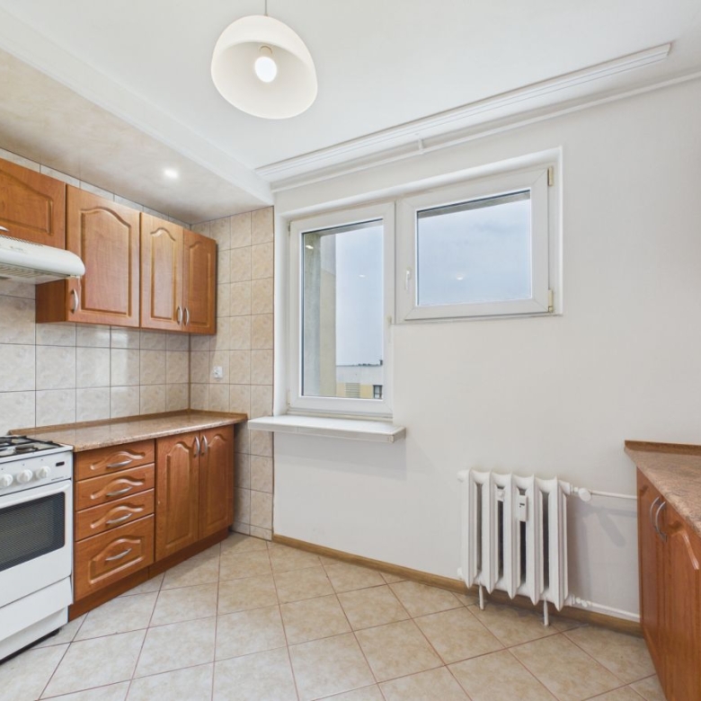 Bright kitchen with wooden cabinets, tiled backsplash, and a white gas stove; window and radiator on the right side—cozy, compact meal-prep space.