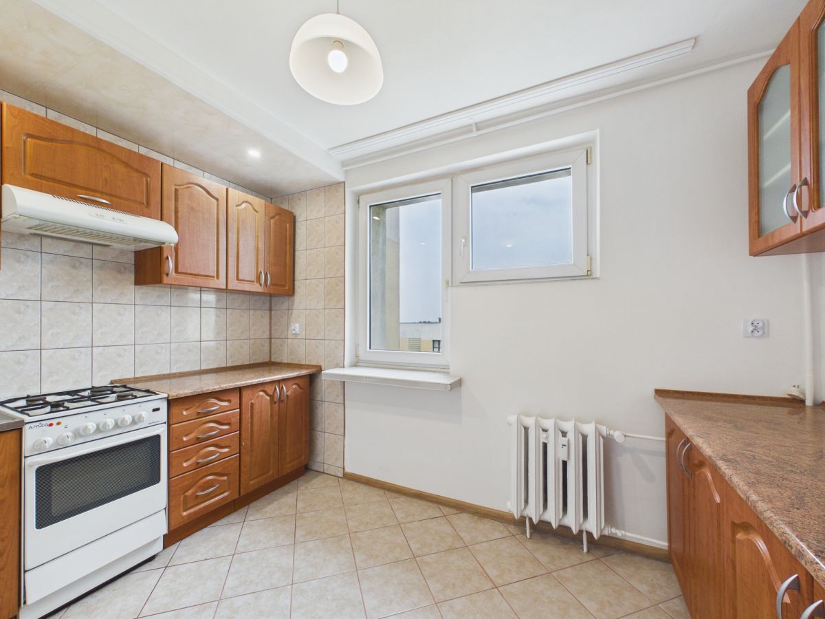 Bright kitchen with wooden cabinets, tiled backsplash, and a white gas stove; window and radiator on the right side—cozy, compact meal-prep space.
