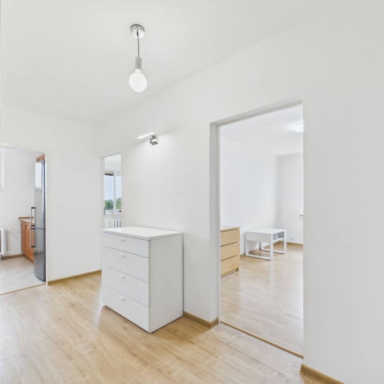 Bright, minimalist hallway with white walls, light wood floors, and a white chest of drawers near the center of the corridor.