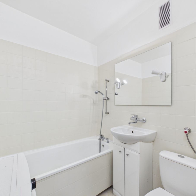 White bathroom with a rectangular tub and handheld shower, a white vanity with sink, a large wall mirror, and a toilet; all fixtures are chrome on off-white tile walls.