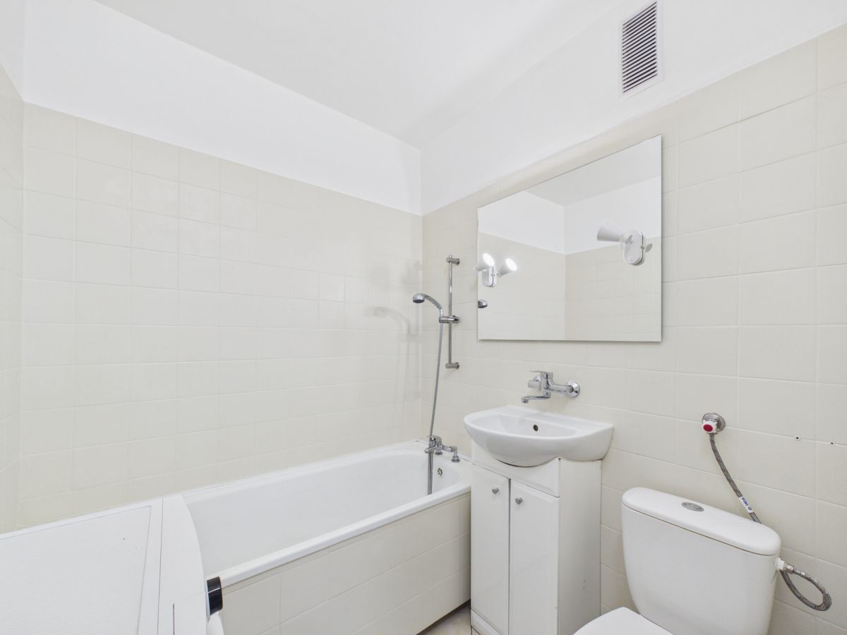 White bathroom with a rectangular tub and handheld shower, a white vanity with sink, a large wall mirror, and a toilet; all fixtures are chrome on off-white tile walls.
