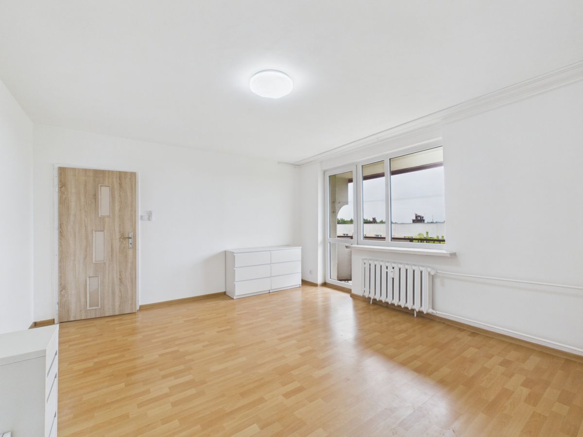 Bright empty living room with light wood flooring, white walls, a wooden door, white dresser, and a large window above a radiator.