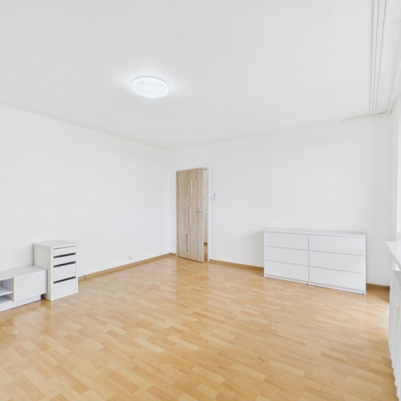 Bright, empty room with white walls and light wood flooring, featuring white storage furniture and a window along the right wall.