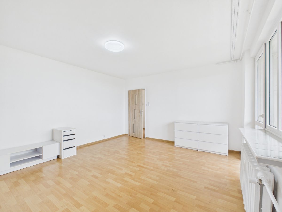 Bright, empty room with white walls and light wood flooring, featuring white storage furniture and a window along the right wall.