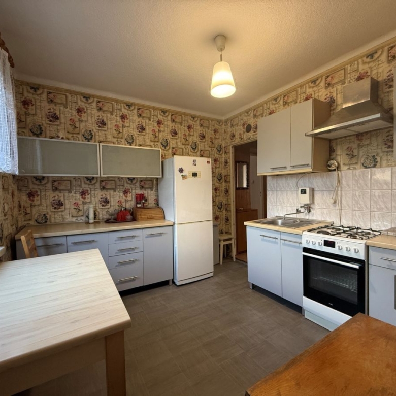 Retro-style kitchen with light gray cabinets, white appliances, and a white refrigerator against floral wallpaper; a window with lace curtains on the left and a wooden table in the foreground.