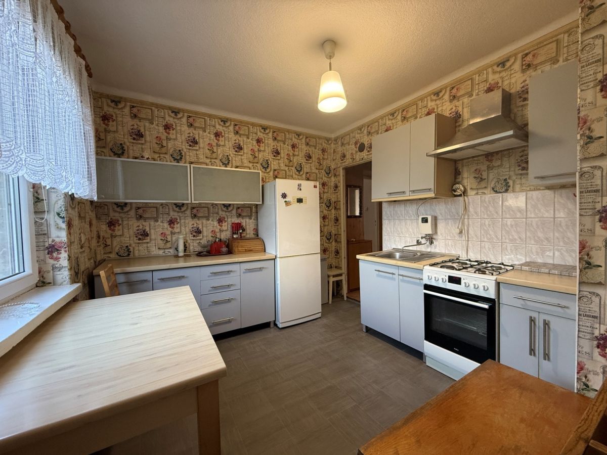 Retro-style kitchen with light gray cabinets, white appliances, and a white refrigerator against floral wallpaper; a window with lace curtains on the left and a wooden table in the foreground.