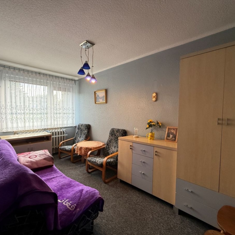 Cozy living room with a purple sofa, two patterned chairs, a small round table, and a wooden cabinet along a light blue wall; lace curtains over a window on the left.