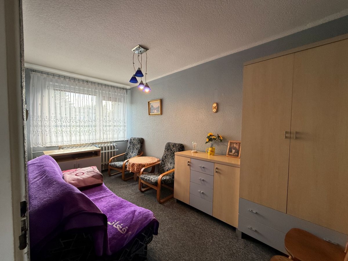Cozy living room with a purple sofa, two patterned chairs, a small round table, and a wooden cabinet along a light blue wall; lace curtains over a window on the left.