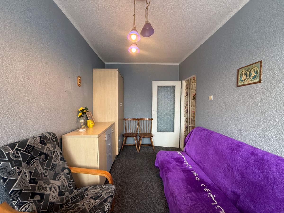 Living room with blue walls, a purple sofa bed along the right and a patterned armchair on the left, facing a wooden dresser and cabinet near the back wall.