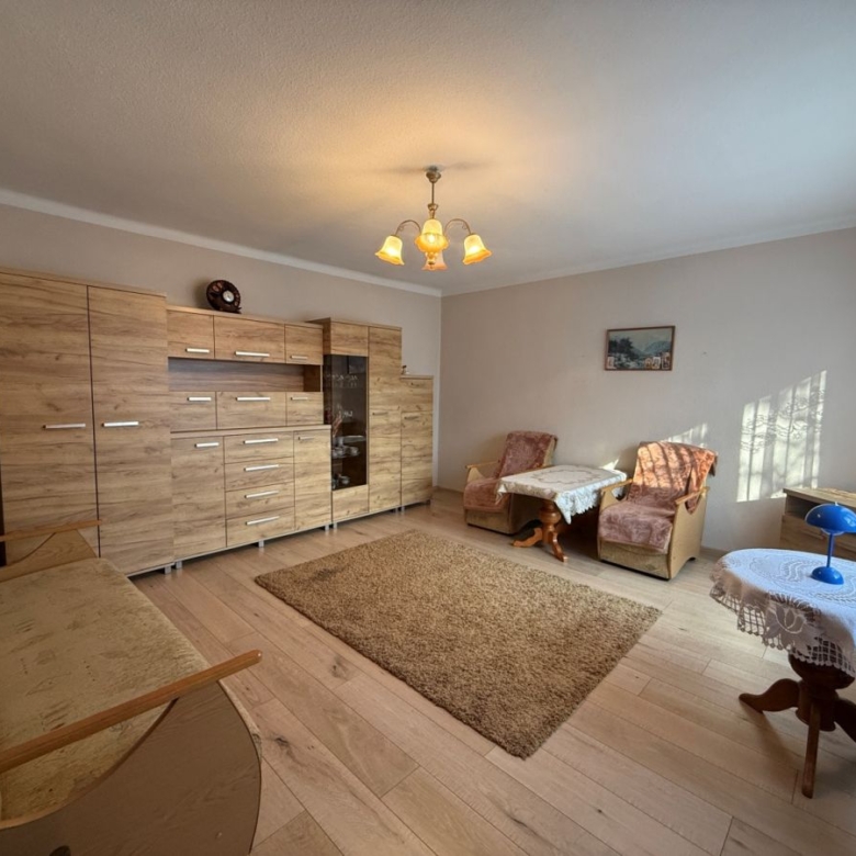 Living room with a large wooden storage wall unit, beige sofa, two pinkish armchairs, a small round table with a lace cloth, and a beige rug underfoot.