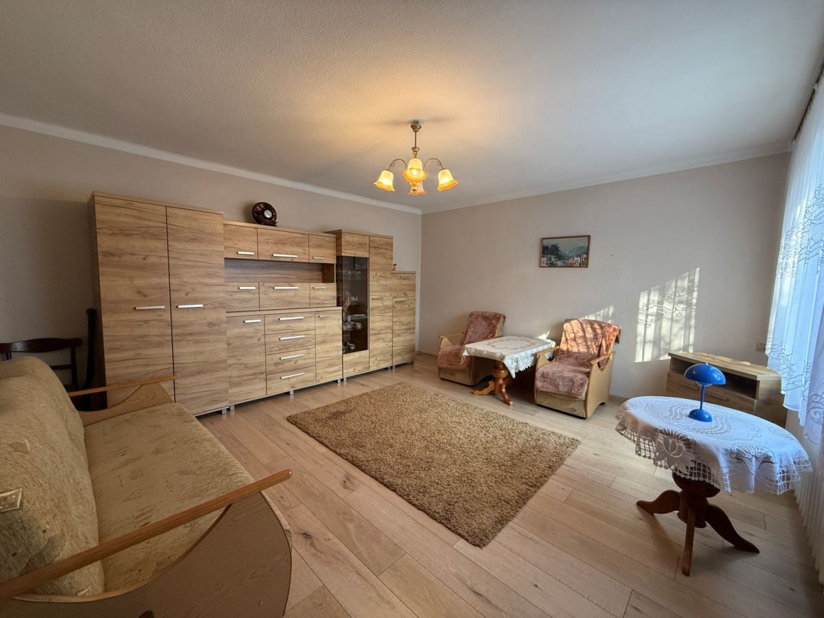 Living room with a large wooden storage wall unit, beige sofa, two pinkish armchairs, a small round table with a lace cloth, and a beige rug underfoot.