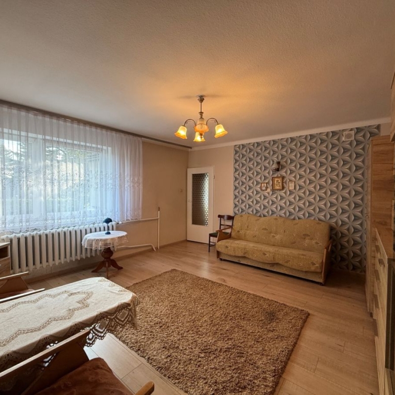 Living room with a beige sofa, wooden furniture, and a large window with sheer lace curtains on the left, radiator beneath the window.