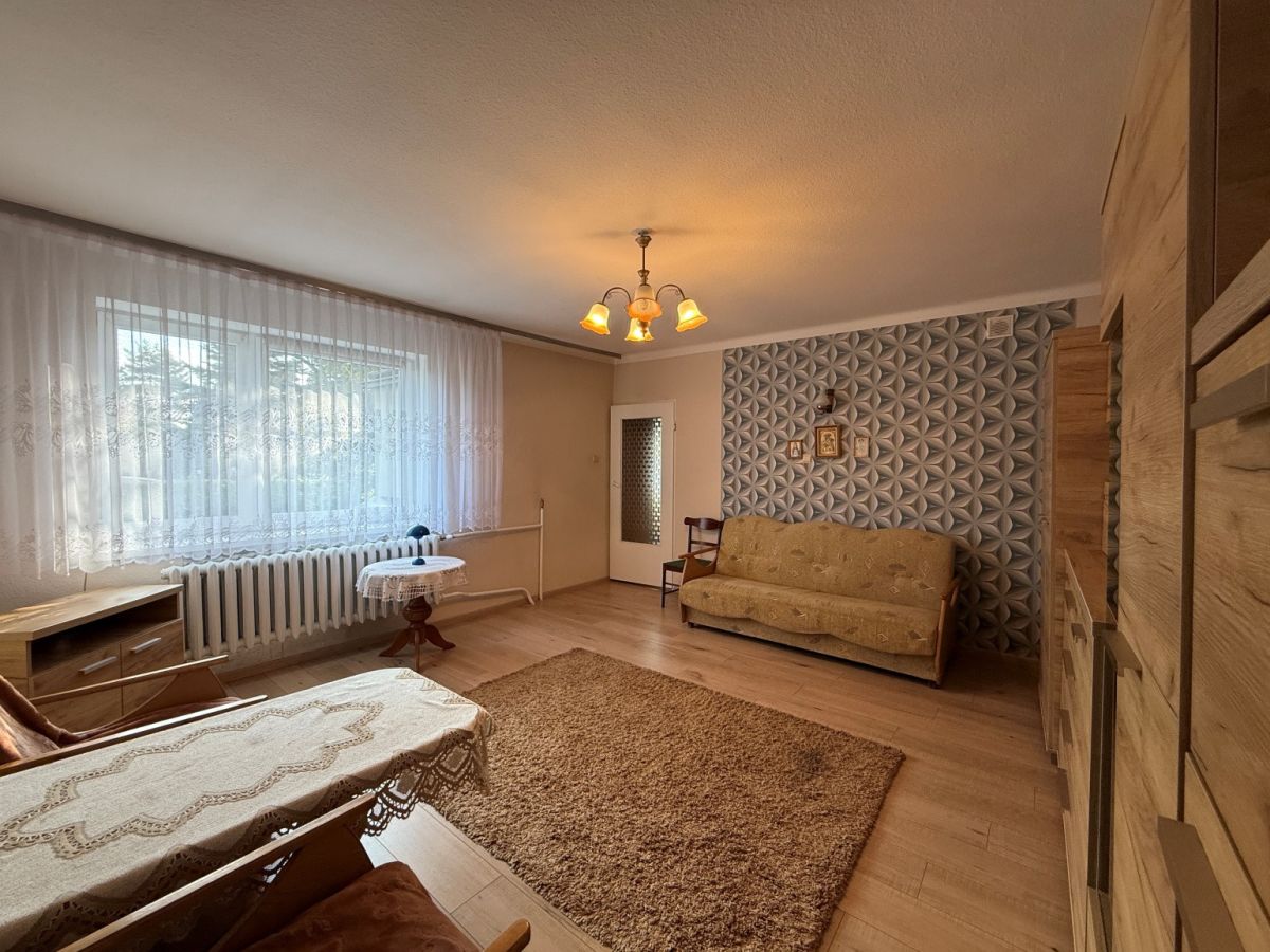 Living room with a beige sofa, wooden furniture, and a large window with sheer lace curtains on the left, radiator beneath the window.