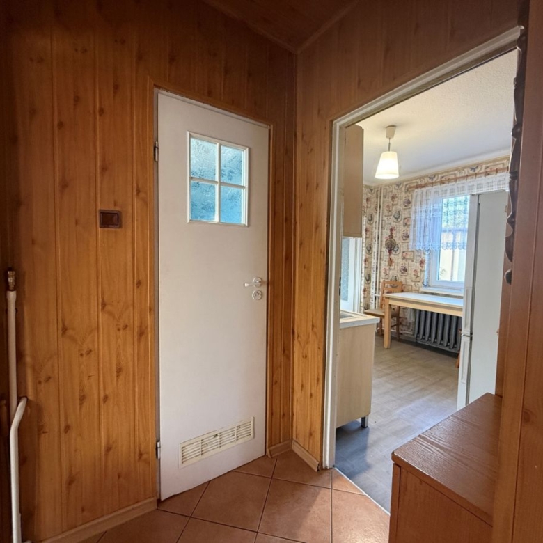 Wood-paneled hallway with a white door, opening into a kitchen/dining area with a window and radiator visible outside the door.