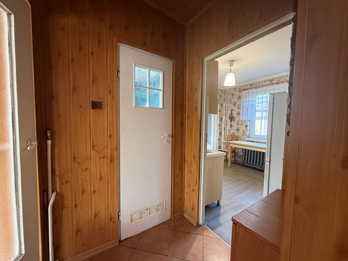 Wood-paneled hallway with a white door, opening into a kitchen/dining area with a window and radiator visible outside the door.
