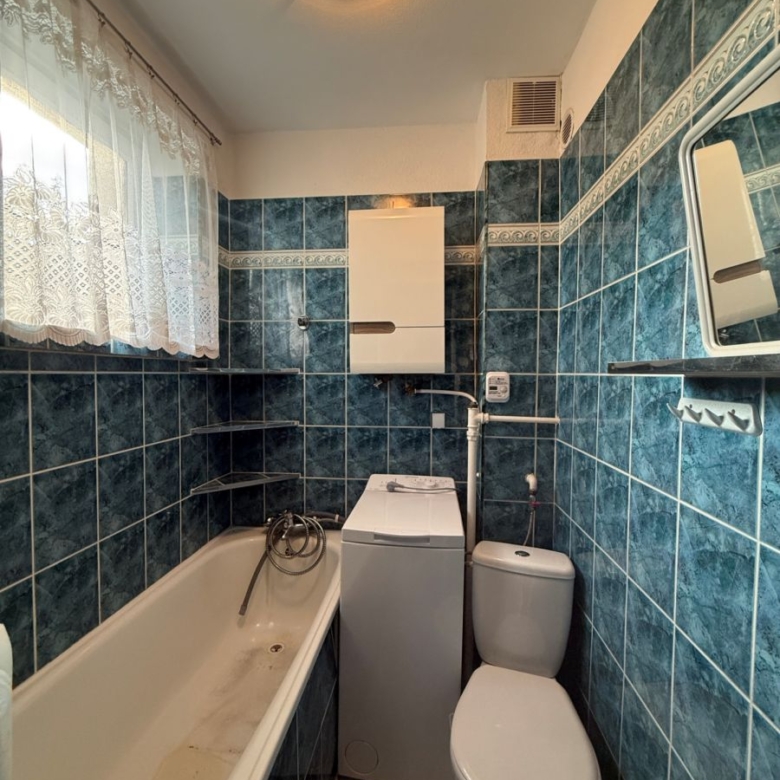 Compact bathroom with blue tiled walls, white bathtub on the left, top-loading washing machine beside the toilet, lace-curtained window.
