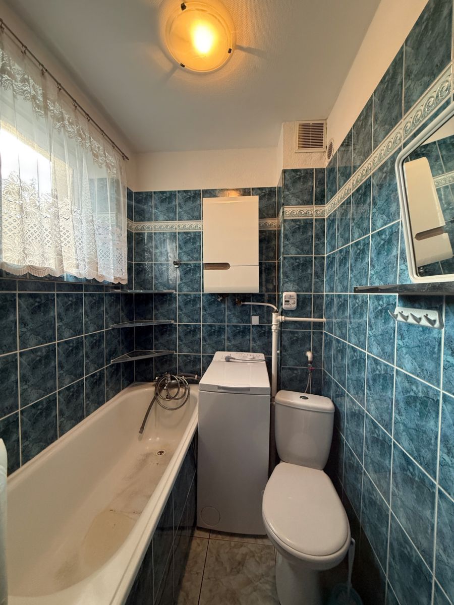 Compact bathroom with blue tiled walls, white bathtub on the left, top-loading washing machine beside the toilet, lace-curtained window.