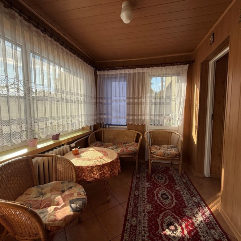 Sunlit enclosed veranda with wicker chairs around a round table and lace curtains along the windows.