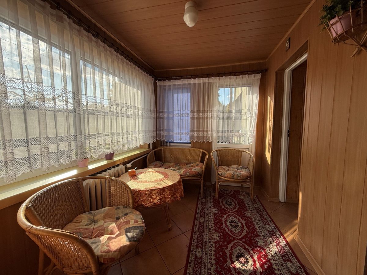 Sunlit enclosed veranda with wicker chairs around a round table and lace curtains along the windows.