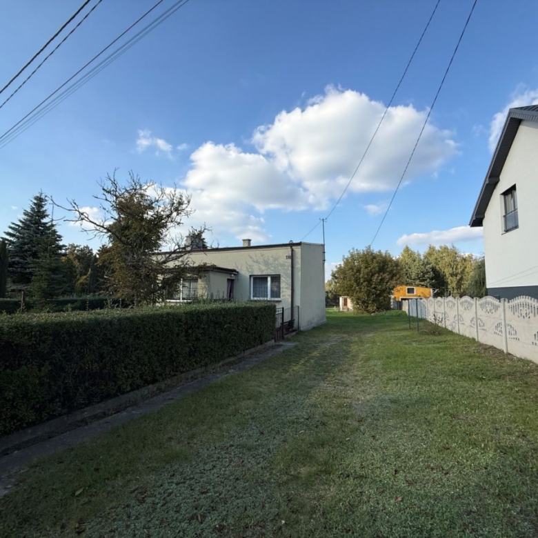 Suburban street scene: small white house center, tall hedge on left, decorative white fence on right, grassy path, blue sky.