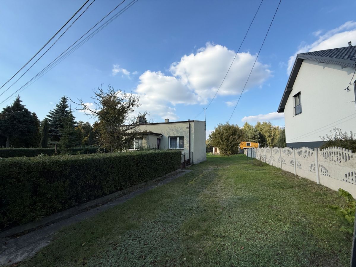 Suburban street scene: small white house center, tall hedge on left, decorative white fence on right, grassy path, blue sky.