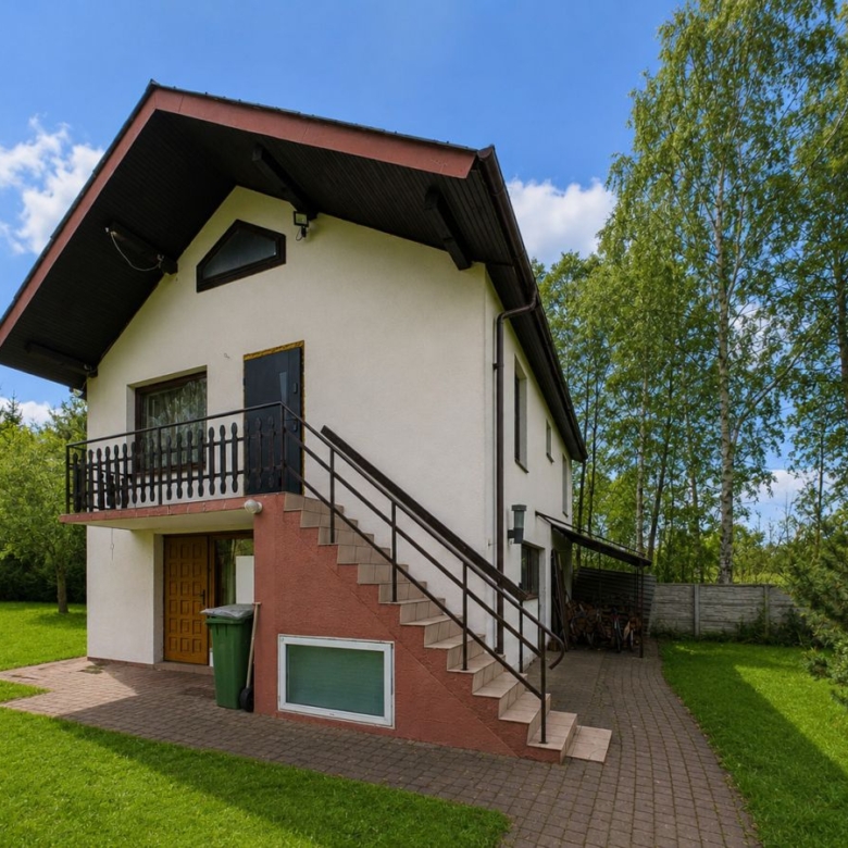 Two-story house with an exterior staircase leading to a small balcony, white walls, and a red base, surrounded by green lawn and trees on a sunny day.