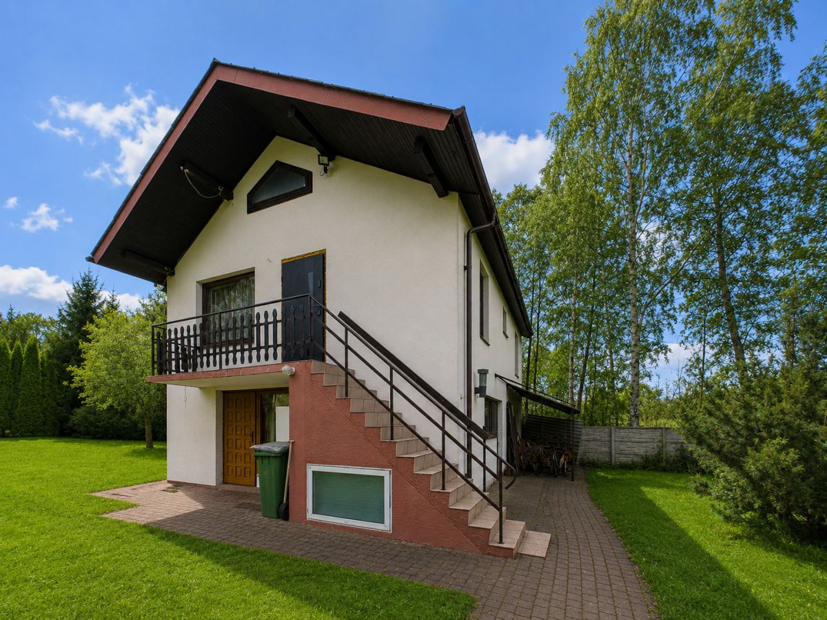 Two-story house with an exterior staircase leading to a small balcony, white walls, and a red base, surrounded by green lawn and trees on a sunny day.