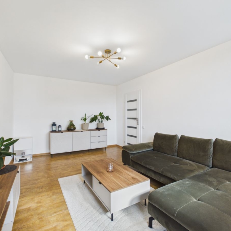 Living room with a dark green sectional sofa, wooden coffee table, and a wall-mounted TV, white walls, and a sideboard with plants.