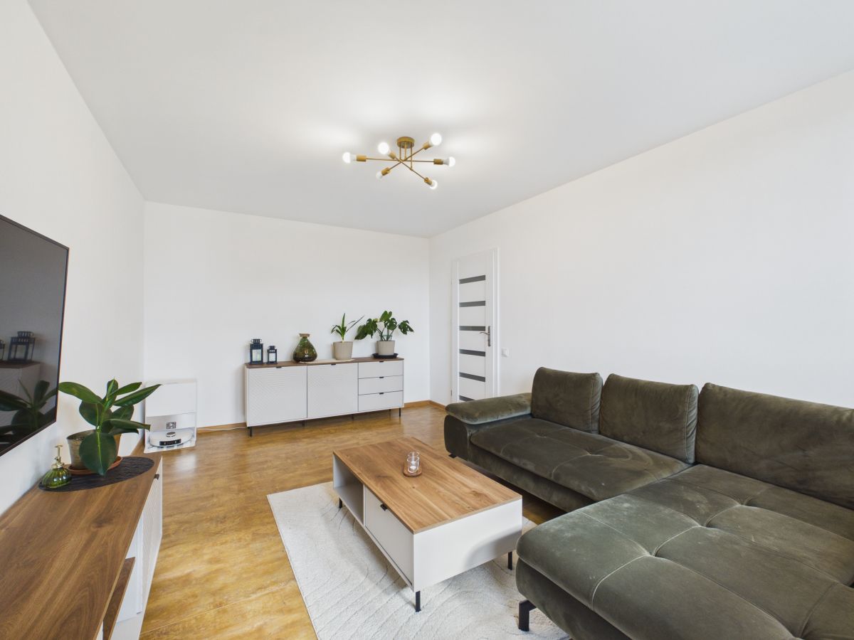 Living room with a dark green sectional sofa, wooden coffee table, and a wall-mounted TV, white walls, and a sideboard with plants.
