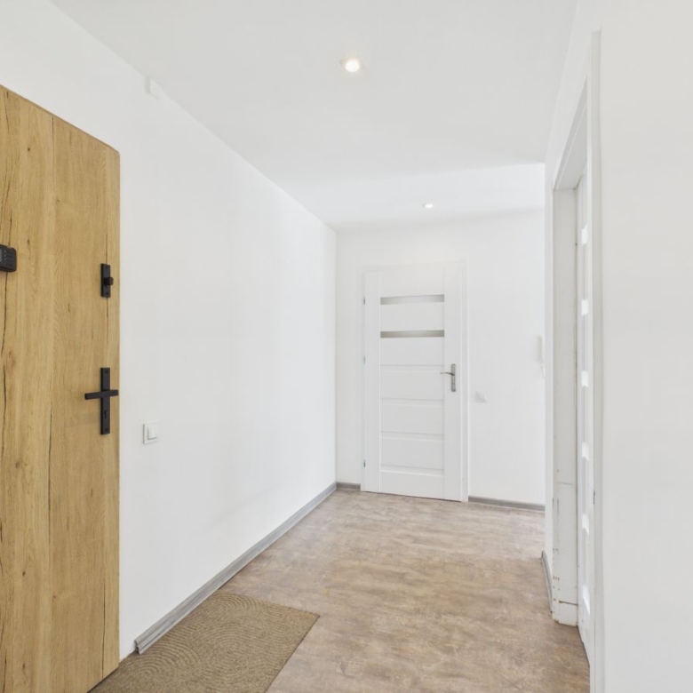 Bright white hallway with a large wood entry door on the left featuring a keypad and a cross handle, and a white panel door at the far end.