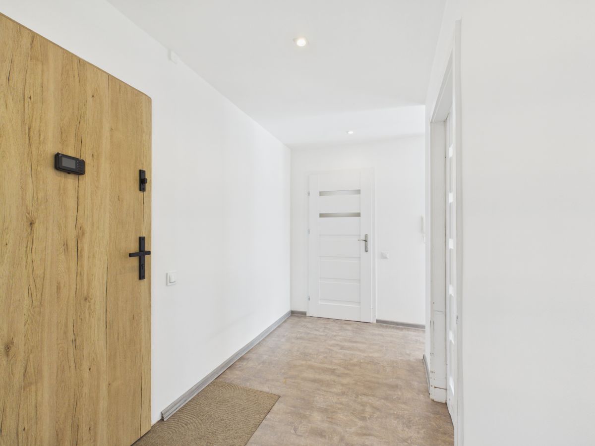 Bright white hallway with a large wood entry door on the left featuring a keypad and a cross handle, and a white panel door at the far end.
