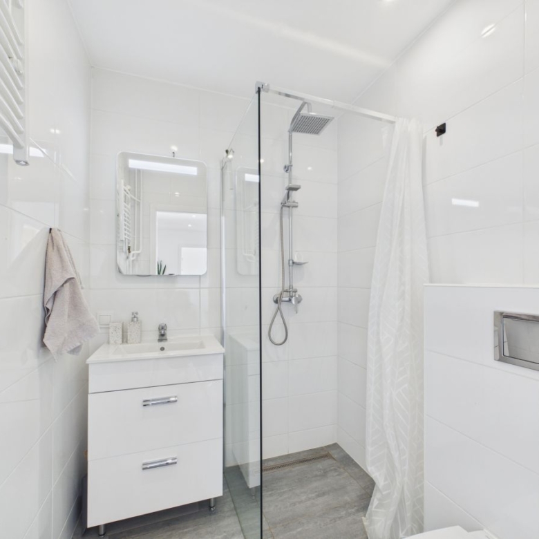 White modern bathroom with a glass shower enclosure, white vanity, and a wall mirror; towel hanging on the left and a light gray floor.