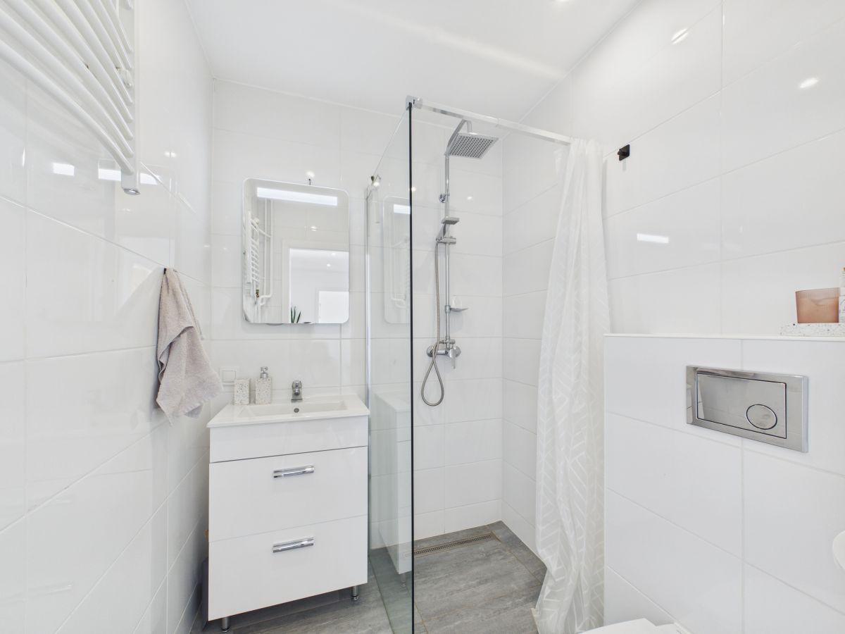White modern bathroom with a glass shower enclosure, white vanity, and a wall mirror; towel hanging on the left and a light gray floor.