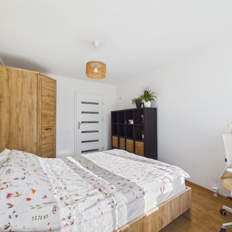 Bright bedroom with a wooden bed, matching wardrobe, and a black cubby storage unit against white walls; a potted plant sits atop the unit.