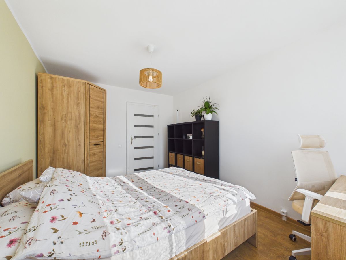 Bright bedroom with a wooden bed, matching wardrobe, and a black cubby storage unit against white walls; a potted plant sits atop the unit.