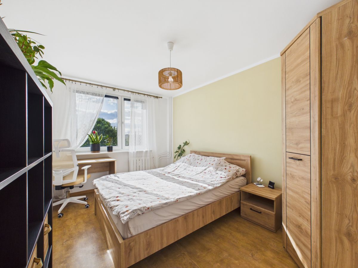 Cozy bedroom with a wooden bed, bedside table, wardrobe, and a desk by a window with potted plants.