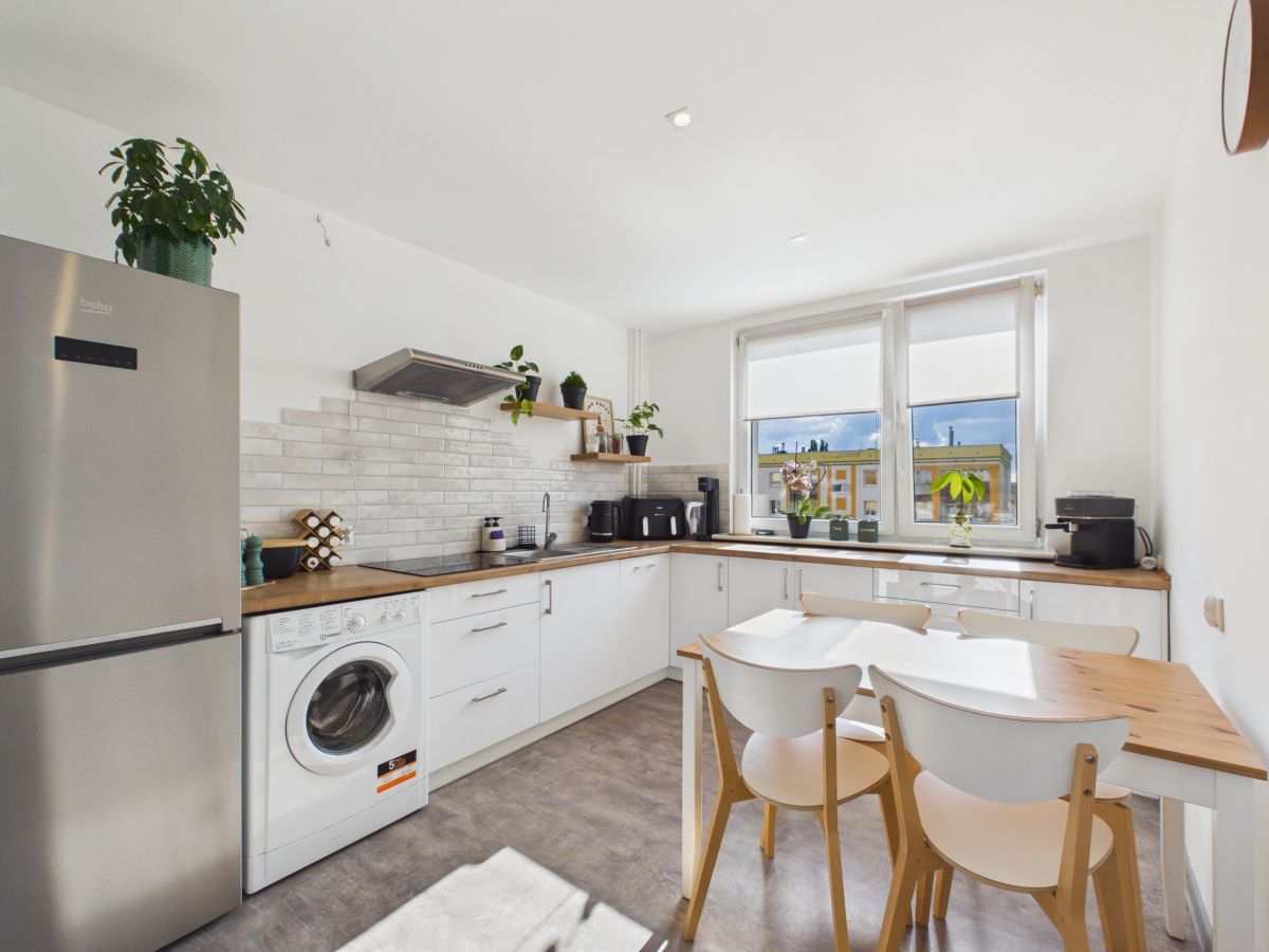 Bright modern kitchen with white cabinets, wooden countertops, and a stainless fridge; a washing machine is tucked under the counter near the left wall.