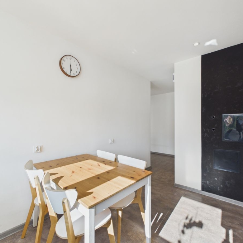 Bright dining area with a wooden table and white chairs, clock on a white wall, and a dark feature wall with a built-in fireplace/oven.