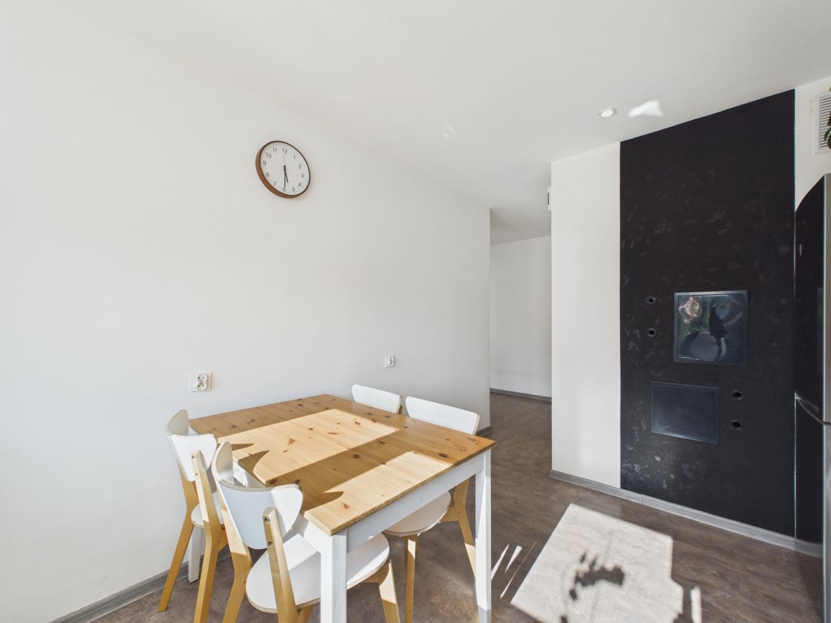 Bright dining area with a wooden table and white chairs, clock on a white wall, and a dark feature wall with a built-in fireplace/oven.