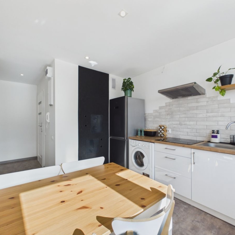 Modern kitchen with a wooden dining table in the foreground, white cabinets, stainless steel fridge, and a washing machine beside a tiled backsplash.