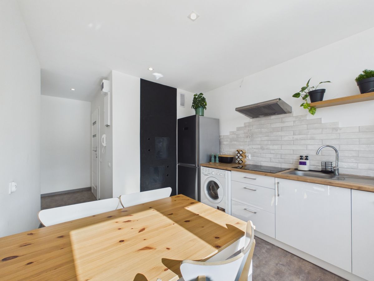 Modern kitchen with a wooden dining table in the foreground, white cabinets, stainless steel fridge, and a washing machine beside a tiled backsplash.