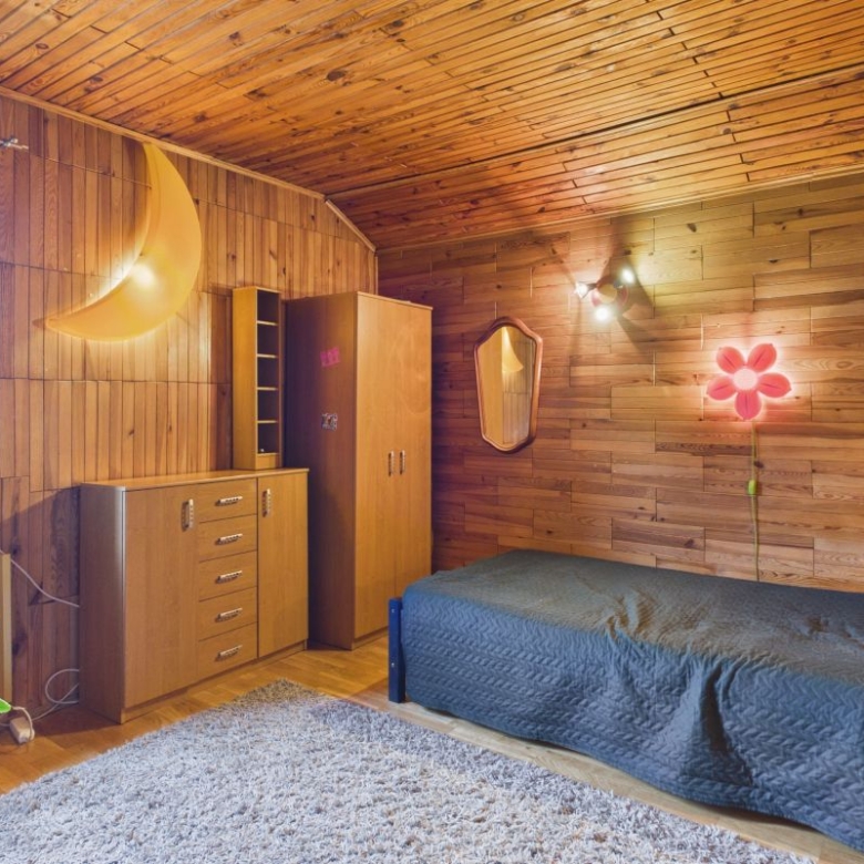 Cozy wood-paneled bedroom with a single bed, desk dresser, and large plant near a window; warm lighting and a crescent moon wall lamp create a vintage vibe.