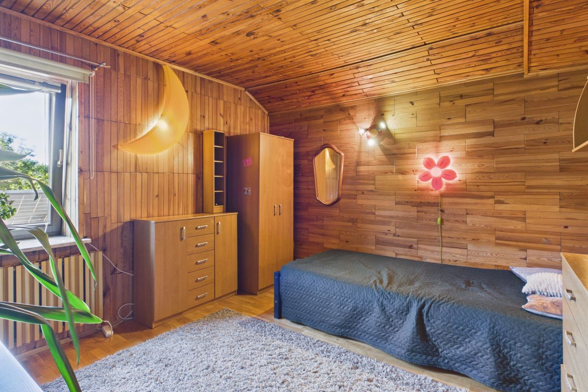 Cozy wood-paneled bedroom with a single bed, desk dresser, and large plant near a window; warm lighting and a crescent moon wall lamp create a vintage vibe.