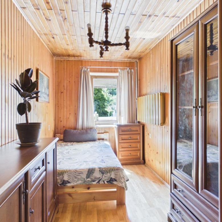 Cozy wood-paneled bedroom with a bed near a window, dresser, and potted plant in warm daylight. In a rustic cabin-style room with wooden walls and ceiling.