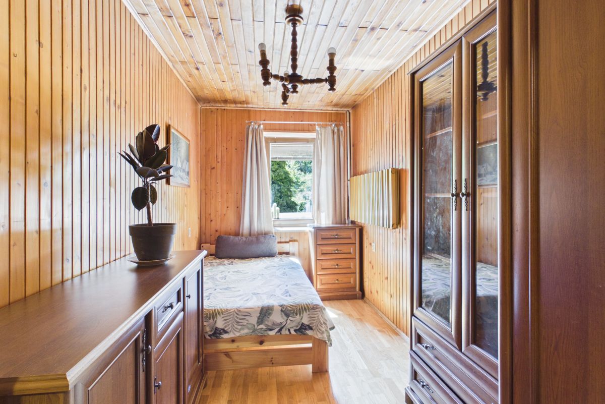 Cozy wood-paneled bedroom with a bed near a window, dresser, and potted plant in warm daylight. In a rustic cabin-style room with wooden walls and ceiling.