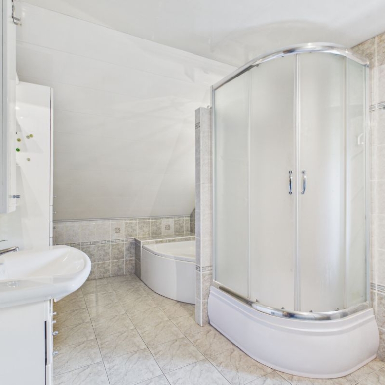 Bright bathroom with curved glass shower enclosure, white tub, and sink; tiled walls and floor with a green mosaic accent near the sink and a window to the right